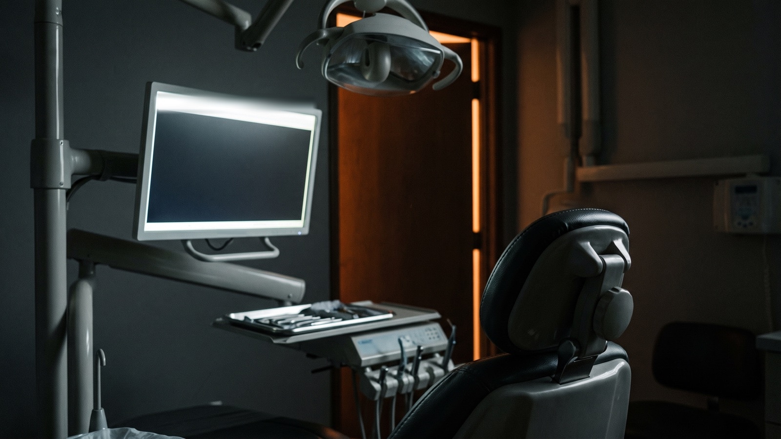 A darkened dental surgery after hours, the dental chair and overhead operatory light in shadow while the practice computer beside the chair is still switched on
